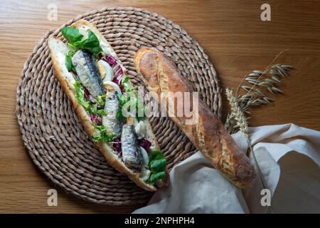 Baguette-Sandwich mit Sardine, Eiern, gemischtem Salat und Mayonnaise, weit geschnitten, serviert auf einer rustikalen Unterlage, auf einem Holztisch mit Backtuch, Top V Stockfoto