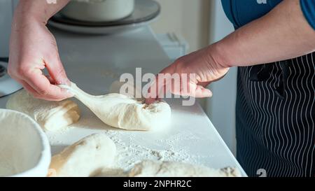 Der Koch streckt und faltet Brotteig auf dem weißen Tisch mit Blick auf das staubige Mehl. Handgemachtes Sauerteigbrot, Hausgebäck bei Tageslicht. Stockfoto