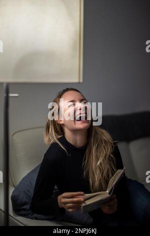 Begeisterter Leser, der lacht. Junge attraktive Frau glückliches positives Lächeln Sitzen Sofa zu Hause Relax lesen Buch Geschichte Neuheit Lachen Humor Witz Stockfoto