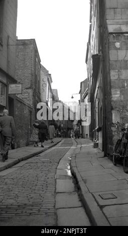 1950er Jahre, historisch, mit Blick vom Boden, aufwärts von den Trümmern, eine enge alte mittelalterliche Kopfsteinpflasterstraße in York, England, Großbritannien. Auf der linken Seite des Bildes ist der Lederladen Cox of Northampton zu sehen, der 1921 von William Cox, einem Schuhfabrikanten und Schuhmacher, gegründet wurde, der in die Domstadt gezogen war. Das Geschäft wurde 93 Jahre lang gehandelt, bevor es 2014 geschlossen wurde. Stockfoto