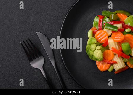Köstliches saftiges Brokkoli-Gemüse, Karotten, Spargelbohnen, Paprika gedünstet auf einer schwarzen Platte auf dunklem Betonhintergrund Stockfoto
