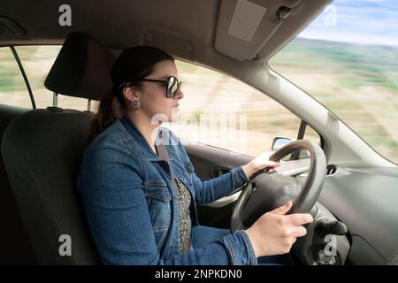 Seitenansicht einer jungen lateinin in Blau mit Brille, die ihr Auto tagsüber mit hoher Geschwindigkeit fährt Stockfoto