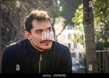 Der junge Mann sitzt auf der Bank im Park in Rom. Ein hübscher Mann im schicken Anzug wartet auf seine Freundin. Stockfoto