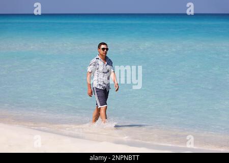 Porträt des Menschen in Sonnenbrille, der am weißen Sand in blauen Ozeanwellen vorbeigeht. Urlaub am tropischen Strand der Karibikinsel Stockfoto