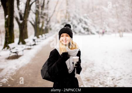 Eine junge Frau steht in einem Winterwunderland, trägt warme Kleidung und trinkt heißen Kaffee zum Mitnehmen Stockfoto