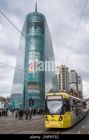 Im Februar 2023 fährt eine gelbliche Straßenbahn auf dem Manchesters Metrolink am National Football Museum vorbei. Stockfoto
