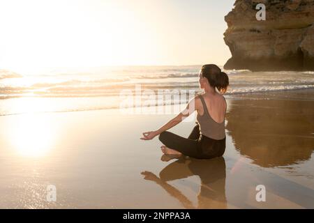 Eine Frau, die in Lotusposition am Strand sitzt, meditiert und Yoga praktiziert. Stockfoto