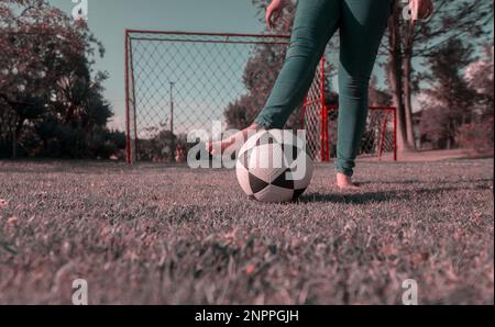 Nahaufnahme der Beine einer Frau in blauer Hose, barfuß mitten im Park und mit einem Fußballball mit einem Fußballtor mit Netzen und Bäumen im Hintergrund Stockfoto