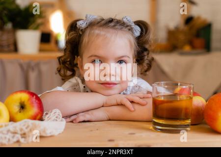 Süßes kleines Mädchen in der Küche zu Hause mit Äpfeln und einem Glas Apfelsaft. Umweltfreundliche Kinderkost. Platz für Text. Hochwertiges Foto Stockfoto