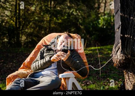 Eine tragbare Solarbatterie hängt an einem Baum. Ein Mann, der in der Natur ruht, mit einem Handy in der Hand. Aufladen des Telefons mit Solarenergie. Stockfoto