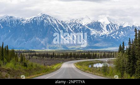 Der Alaska Highway fährt im Frühling in die Stadt Haines Junction mit epischen, riesigen Bergen in weiter Ferne und einer atemberaubenden Panoramastraße. Stockfoto