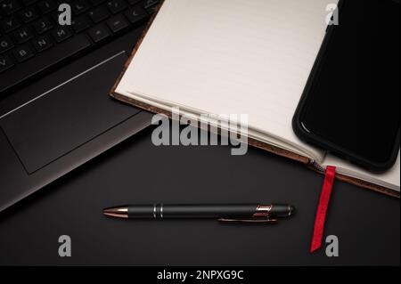 Ein mit Lesezeichen versehenes Notebook, Mobiltelefon, Stift und Laptop auf einem schwarzen Desktop, das als Vorbereitung für kreatives Schreiben das Gefühl eines Home Office vermittelt. Stockfoto