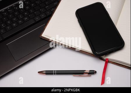 Ein mit Lesezeichen versehenes Notebook, Mobiltelefon, Stift und Laptop auf einem weißen Desktop-PC, das als Vorbereitung auf kreatives Schreiben das Gefühl eines Home Office vermittelt. Stockfoto