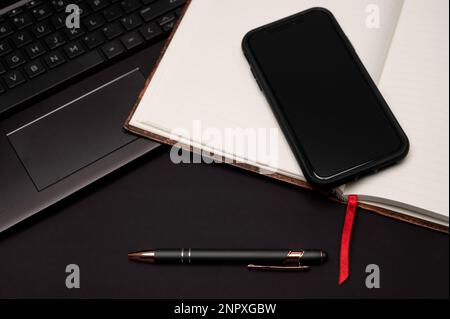 Ein mit Lesezeichen versehenes Notebook, Mobiltelefon, Stift und Laptop auf einem schwarzen Desktop, das als Vorbereitung für kreatives Schreiben das Gefühl eines Home Office vermittelt. Stockfoto