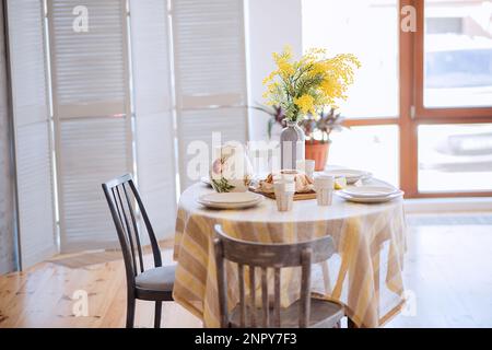 Frühling, Ostern-Interieur im skandinavischen Stil. Rustikales Wohnzimmer mit einem hellen Kleiderschrank, gelbe Mimosa in Vase. Der Tisch ist festlich mit Keramik gedeckt Stockfoto