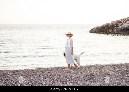Schwangere zukünftige Mutter, die mit einem goldenen Retriever am Strand entlang spaziert. Eine Millennials-Frau mit weißem Hut hält den Hund an der Leine. Konz Stockfoto