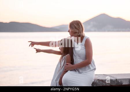 Schwangere Mutter der Millennials sitzt mit ihrer Tochter bei Sonnenuntergang am Meer. Eine Frau mittleren Alters umarmt ein kleines Mädchen, zeigt mit dem Zeigefinger auf den Berg. Das j Stockfoto