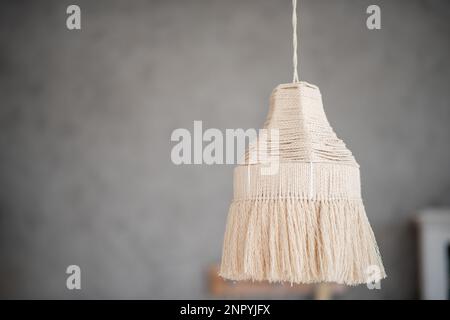 Die Makrame-Lampe hängt auf einem grauen, einfarbigen Hintergrund. Handgemachtes Hobby. Dekoration im Boho-Stil. Skandinavische Wohnungseinrichtung. Trendige Designdetails. Beige Stockfoto