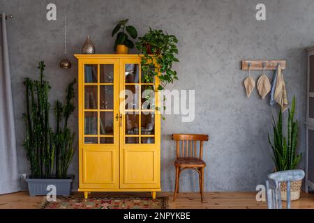 Dunkelgelbes Bücherregal aus Holz an grauer Betonwand. Um grüne Töpfe mit Hausblumen. Skandinavisches Apartment im Loftstil, rustikales Design. Makrame Stockfoto