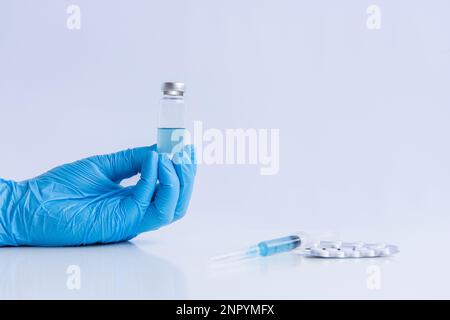 Auf weißem isoliertem Hintergrund halten die Hände des Arztes in medizinischen blauen Handschuhen eine Ampulle Impfstoff gegen Virus Coronovirus. Spritze mit Gegenmittel, Schmerztabletten Stockfoto