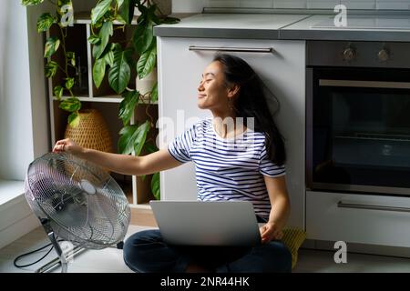 Fröhliche, lächelnde Freiberuflerin in Asien, die neben einem elektrischen Ventilator sitzt und von zu Hause aus in Hitzewelle arbeitet Stockfoto