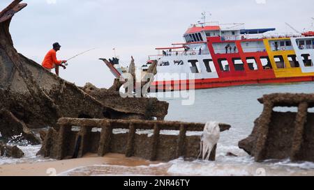 Eine vorbeifahrende Fähre und Ein Fischer stehen auf Einem versunkenen Schiffswrack am Strand in Muntok Town am Nachmittag Stockfoto