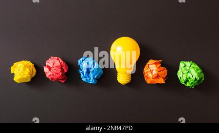 Bunte zerknitterte Papierkugel mit gelber Glühbirne vor schwarzem Hintergrund Stockfoto