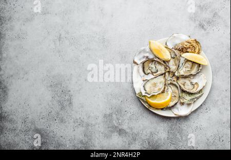 Nahaufnahme eines halben Dutzend frischer Austern und Schalen mit Zitronenkeilen auf einem Teller, Draufsicht, grauer rustikaler Betonhintergrund, Platz für Text. Stockfoto