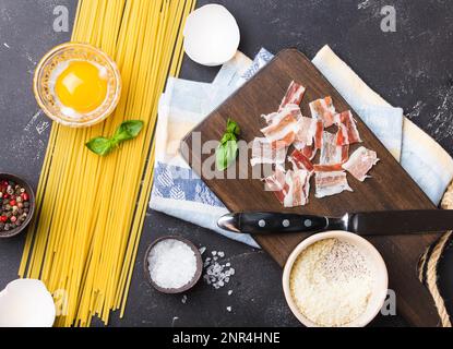 Zutaten zum Kochen traditioneller italienischer Nudelgerichte Spaghetti Carbonara, rohes Eigelb, Parmesankäse, Speck auf Holzbrett auf schwarzem rustikalem Stein Stockfoto