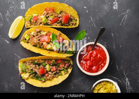 Blick von oben auf mexikanische Tacos mit Fleisch, Gemüse in gelber Maistortilla serviert mit Limette, Salsa und Guacamole Saucen auf schwarzem rustikalem Stein Stockfoto