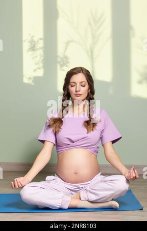 Eine schwangere Frau mit langen braunen Haaren setzt sich quer, meditiert und übt Yoga für körperliche Fitness und Wohlbefinden während der Schwangerschaft. Auf dem Ba Stockfoto