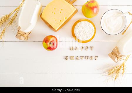 Verschiedene Arten von Milchprodukten in Schüsseln, Flaschen auf weißem Hintergrund. Draufsicht. Flach verlegt. Holzbriefe mit dem Text shavuot Stockfoto