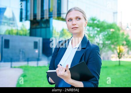 Geschäftsfrau im Anzug ist im Stadtzentrum vor einem Gebäude im Hintergrund, schreibt, schreibt, macht Notizen, führt Stift und Notizbuch, führt Brainstorming, kreiert Ideen. Stockfoto