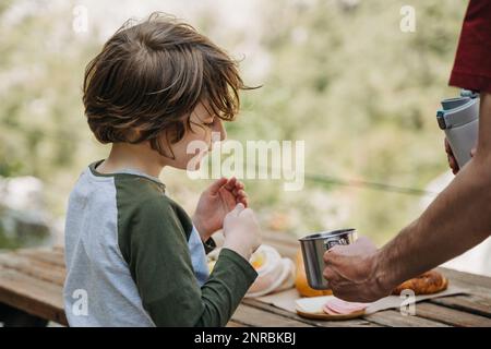 Vater Vater gießt heißen Kaffee-Tee aus Thermoskannen in den Becher bei einem Familienpicknick in den Bergen. Das Kind der Kinderschule sieht zu, wie sein Vater die füllt Stockfoto