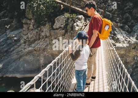 Vater und Sohn blicken beim Wandern weit von der Seilbrücke in die Berge. Lässig gekleidete Touristen Kinderschultjunge und sein Vater mit gelb Stockfoto