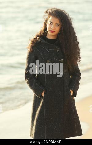 Eine Frau in einem Mantel, die am Strand steht und wegschaut Stockfoto