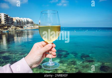 Aus nächster Nähe sehen Sie eine Frau, die ein Glas Weißwein hält, und die wunderschöne Insel Malta im Küstenstädtchen Gozo Marsalforn im Hintergrund mit blauem Himmel. Stockfoto