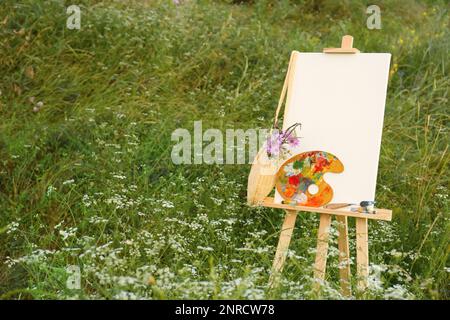 Hölzerne Staffelei mit leerer Leinwand, Malmaschinen und Blumen auf der Wiese. Platz für Text Stockfoto