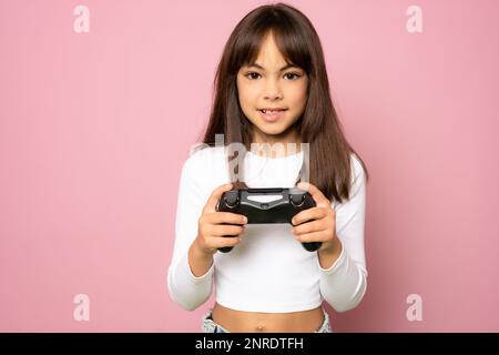 Porträt eines kleinen Mädchens, das gestreiftes T-Shirt trägt, den roten Gamepad-Joystick in den Händen hält, mit optimistischem Ausdruck in die Kamera schaut und das Spiel genießt. In Stockfoto