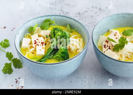 Vegane Currylaksa mit Reisnudeln, Brokkoli und Tofu in blauen Schüsseln, grauer Hintergrund. Stockfoto