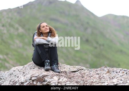 Glücklicher Wanderer, der auf einem Felsen sitzt und die Aussicht auf den Berg erwägt Stockfoto