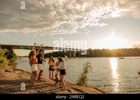 Männliche und weibliche Multirassenfreunde tanzen bei Sonnenuntergang auf Felsen nahe dem Meer Stockfoto