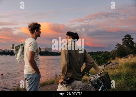 Seitenansicht eines jungen Mannes, der während des Sonnenuntergangs mit einem männlichen Freund mit dem Fahrrad spricht Stockfoto