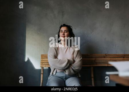 Selbstbewusste Geschäftsfrau mit gekreuzten Armen, die im Büro auf einer Bank sitzt Stockfoto