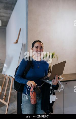 Porträt einer glücklichen Geschäftsfrau mit Laptop- und Handyladegerät, das am Arbeitsplatz steht Stockfoto