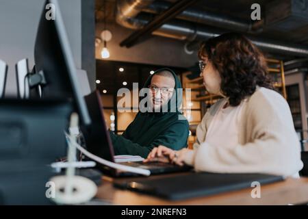 Junger männlicher Hacker, der mit einer Kollegin am Schreibtisch in einem kreativen Arbeitsumfeld spricht Stockfoto