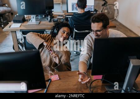 Lächelnder männlicher Hacker, der mit einer Kollegin am Schreibtisch im Kreativbüro spricht Stockfoto