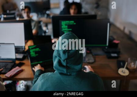 Rückansicht eines jungen männlichen Hackers mit Haubencodierung am Computer am Schreibtisch am kreativen Arbeitsplatz Stockfoto