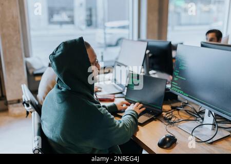 Junger männlicher Hacker, der ein Notebook verwendet, während er am Schreibtisch eines Startup-Unternehmens am Computer codiert Stockfoto