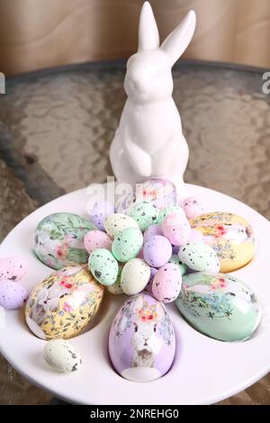 Fotoweißes dekoratives Kaninchen und Eier in verschiedenen Farben und Größen liegen auf einem speziellen Teller für die Eier auf einem Glastisch Stockfoto